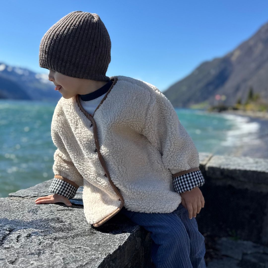 Kind in warmer Kleidung mit Teddyjacke und Mütze am Seeufer in den Bergen.